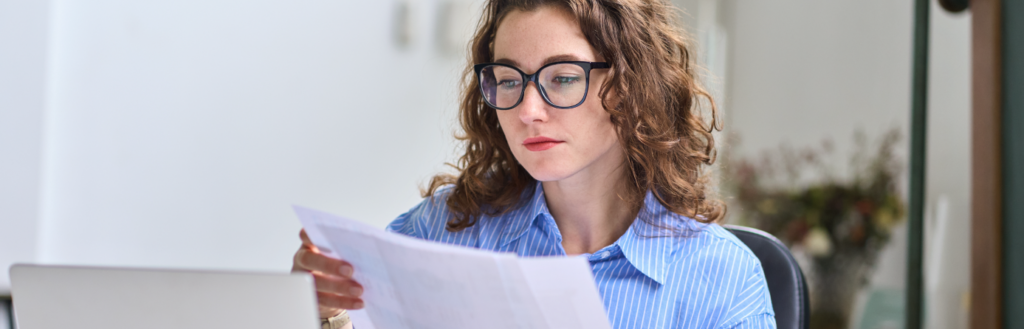 woman wearing glasses while reading paper documents in front of her laptop