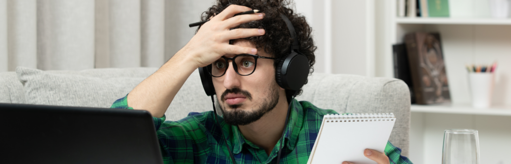 man sitting in front of a laptop while using one hand to hold a pad of paper and the other to frustratingly hold his head